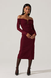 A woman is wearing an elegant, off-the-shoulder, long-sleeved dress in a deep burgundy color. The dress features a ruched design across the bodice and skirt, with a keyhole cutout at the center of the neckline. She accessorizes with large, gold drop earrings and black sheer stockings paired with black pointed-toe heels.